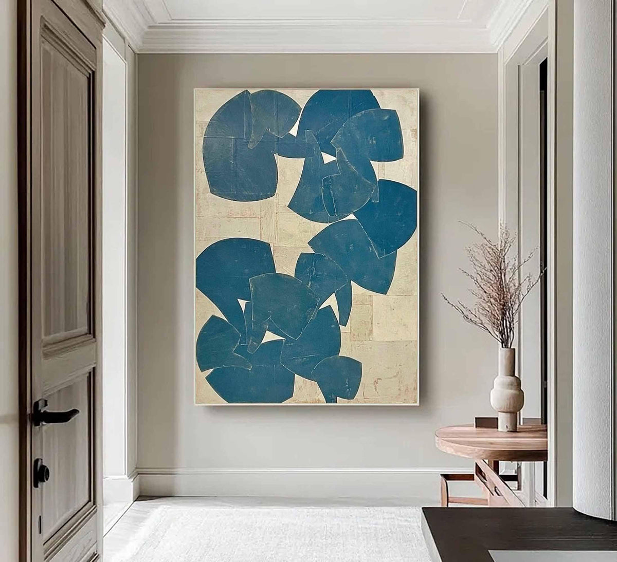 May include: Large abstract painting with blue shapes on a beige background. The artwork is displayed in a hallway with a white rug and a wooden table with a vase. The painting's shapes are irregular and layered.