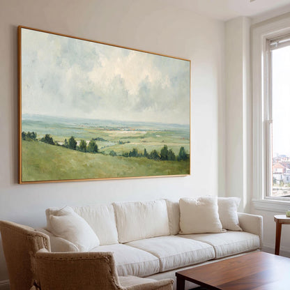 May include: A large landscape painting with a gold frame depicts a serene countryside scene. The artwork features rolling green hills, trees, and a cloudy sky with soft, muted colors. The painting hangs above a white sofa in a well-lit living room.