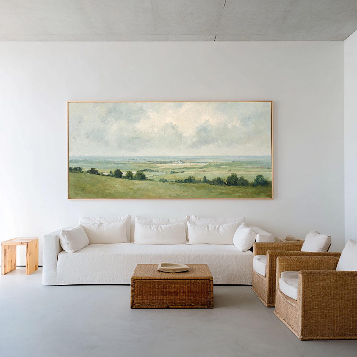 May include: A large landscape painting with a gold frame hangs above a white sofa. The painting depicts a green field, trees, and a cloudy sky. A woven coffee table and two wicker chairs with white cushions complete the living room setting.