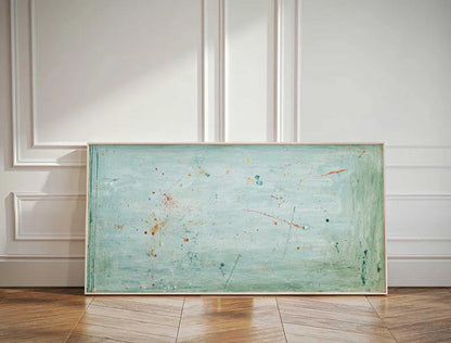 May include: Abstract art piece with a light blue and green background, accented with orange and dark blue paint splatters. The artwork is framed in a light-colored wooden frame and rests against a white wall and wooden floor.