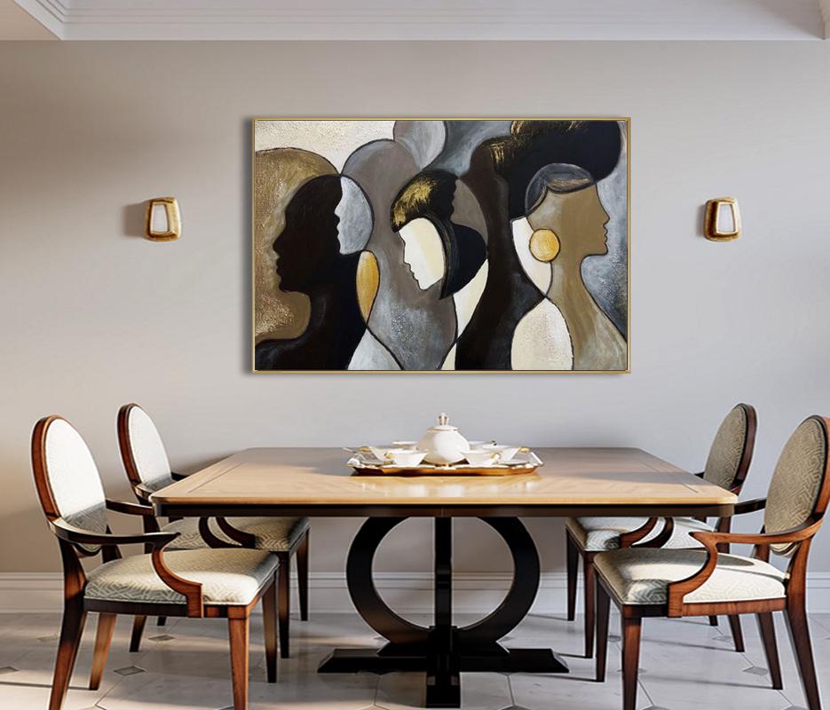 May include: An abstract painting in a gold frame depicts three stylized female profiles in shades of black, white, and gold. The artwork hangs above a wooden dining table with chairs. The room has neutral-toned walls and gold sconces.