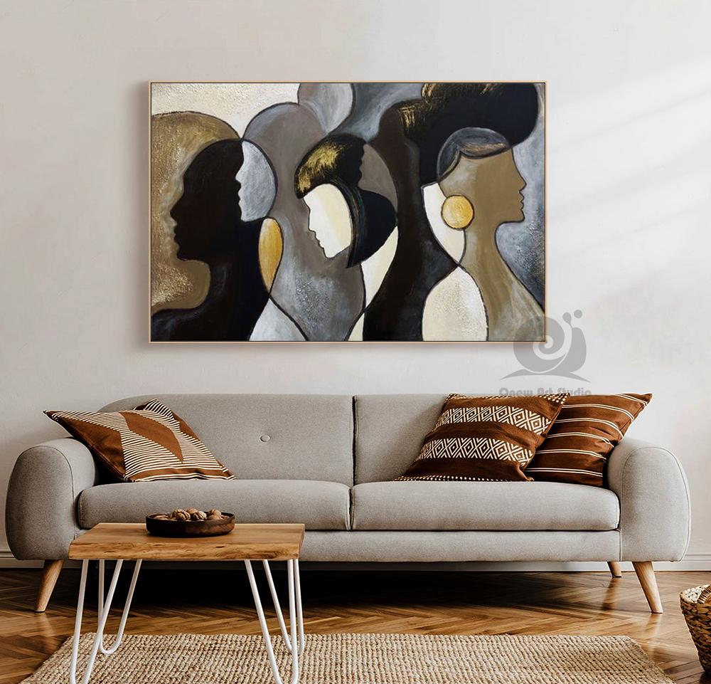 May include: Abstract painting featuring four stylized female profiles in shades of black, gold, and gray. The artwork is framed and hangs above a gray sofa with decorative pillows. The painting's style is modern and minimalist.