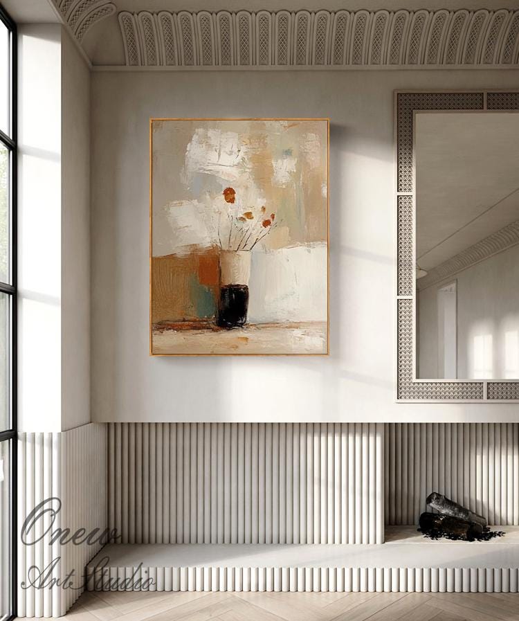 May include: Abstract painting in a gold frame featuring a vase with flowers. The artwork uses a palette of neutral colors, including beige, brown, and white. The vase is black and the flowers are orange. The painting is displayed in a modern interior setting.