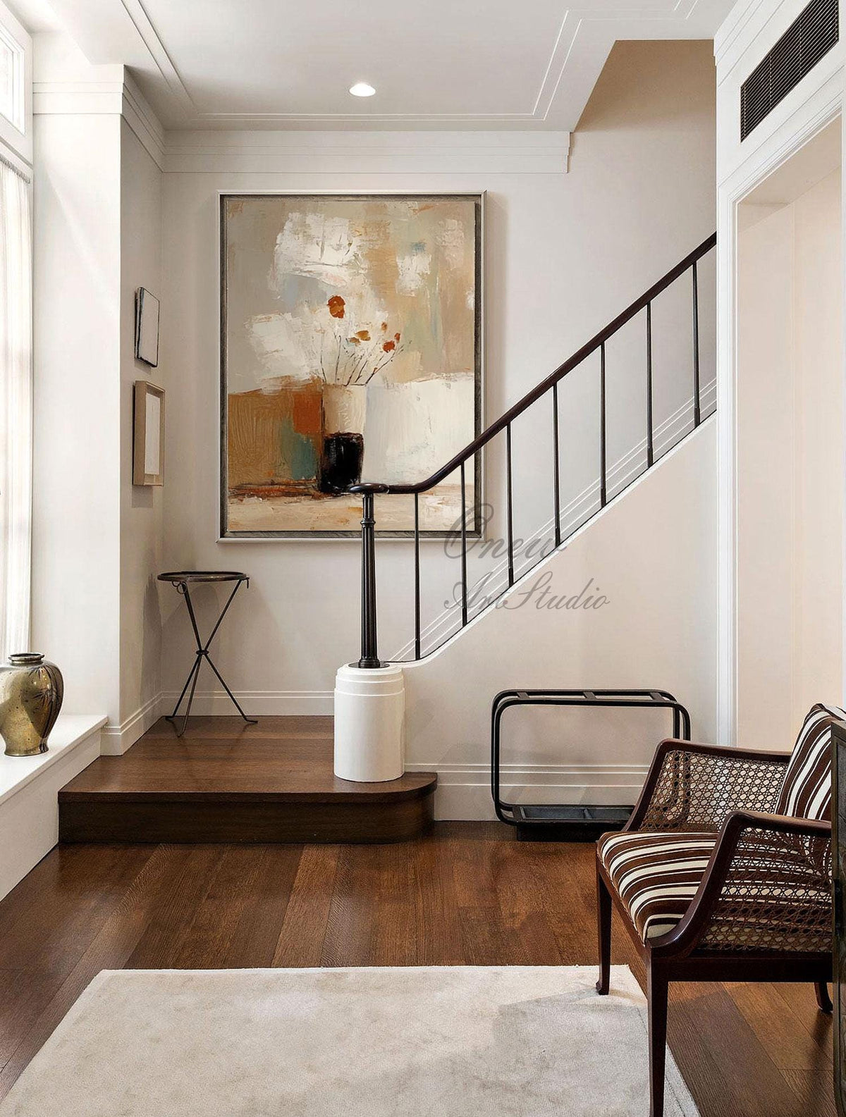 May include: An interior shot of a home with a large abstract painting in neutral tones. A staircase with a dark wood banister and a striped chair are visible. The floor is dark wood, and a light rug is on the floor.