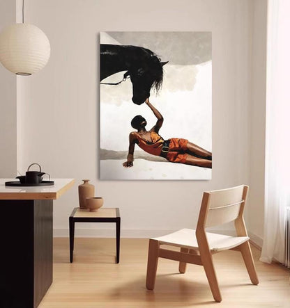 May include: A canvas print depicts a person in an orange outfit reaching towards a black horse's head. The artwork has a neutral background and is displayed in a room with a modern aesthetic, including a wooden chair and side table.