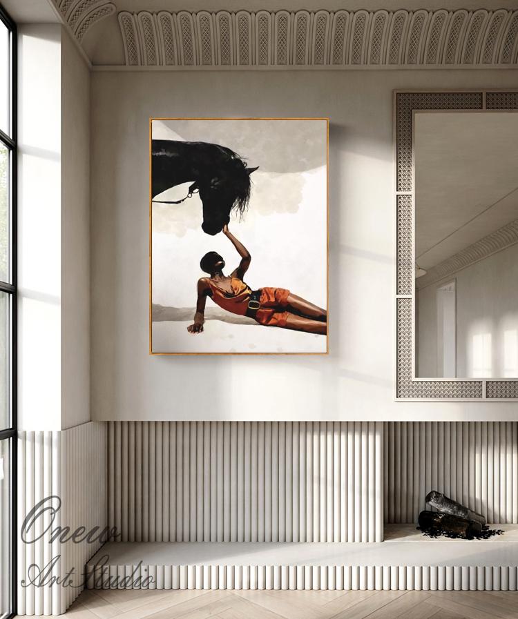 May include: An art print featuring a black horse and a person in an orange outfit. The person is lying down, reaching up to touch the horse's mane. The artwork is framed in gold and hangs on a white wall.