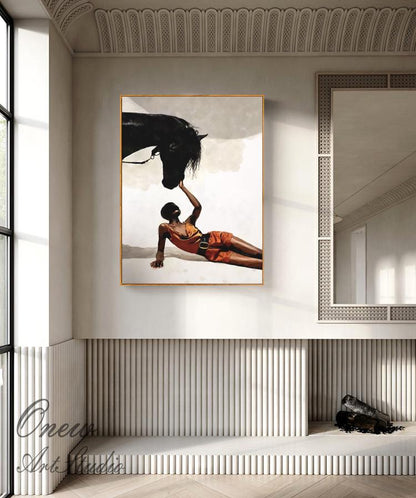 May include: An art print featuring a black horse and a person in an orange outfit. The person is lying down, reaching up to touch the horse's mane. The artwork is framed in gold and hangs on a white wall.