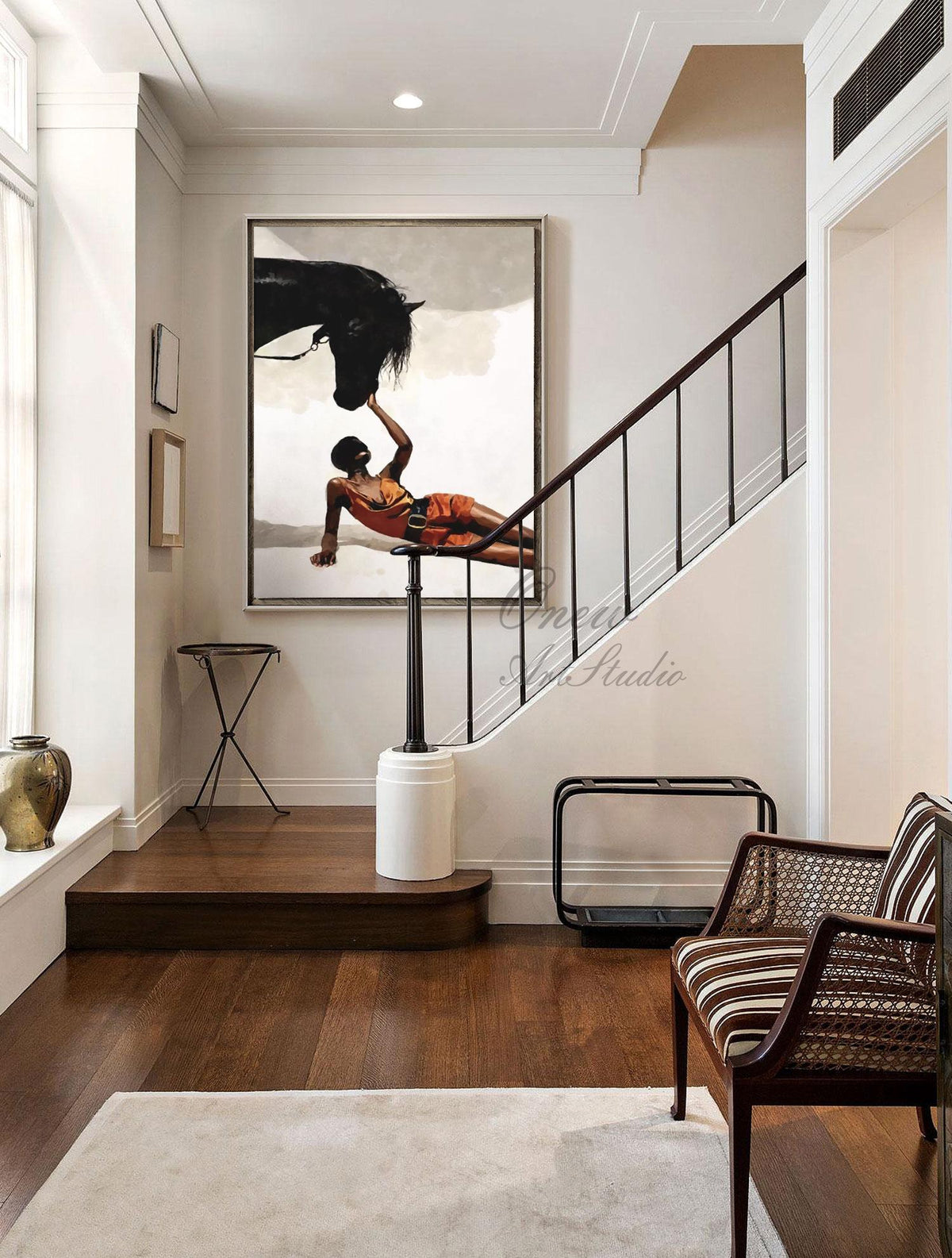 May include: An oil painting depicts a black horse and a person in orange attire. The person is lying down, reaching towards the horse's head. The artwork hangs on a white wall in a well-lit hallway with a staircase and a decorative chair.
