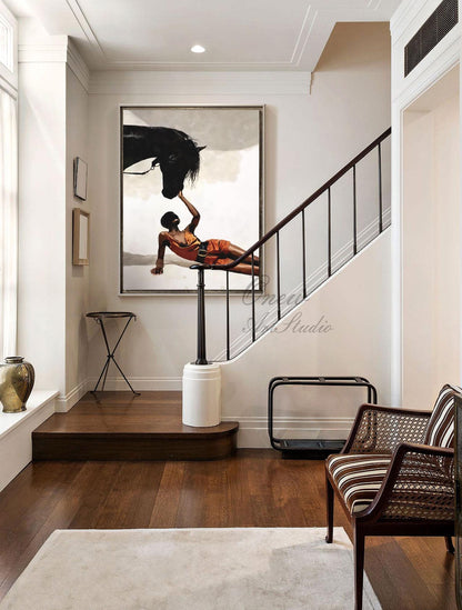 May include: An oil painting depicts a black horse and a person in orange attire. The person is lying down, reaching towards the horse's head. The artwork hangs on a white wall in a well-lit hallway with a staircase and a decorative chair.
