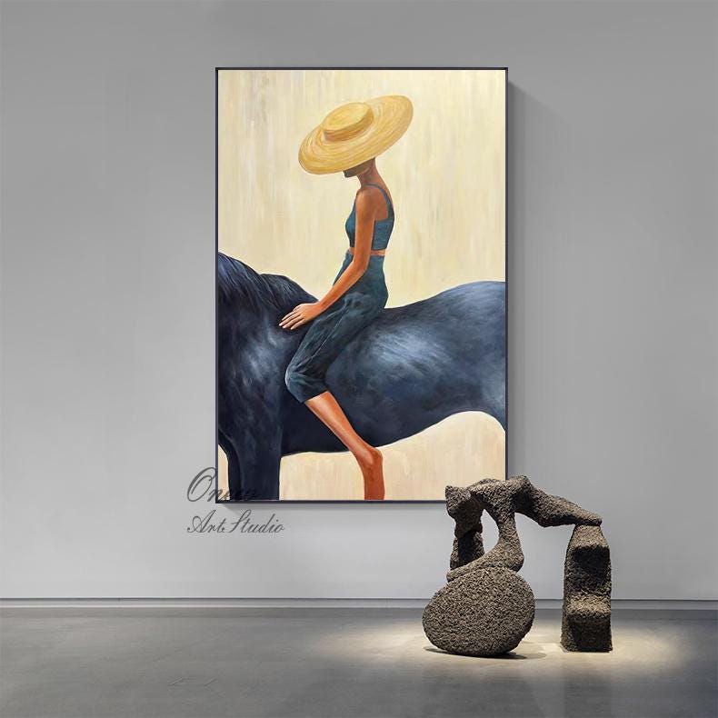May include: An oil painting depicts a person riding a dark blue horse. The person wears a yellow hat and dark teal clothing. The background is a light beige color. The painting is framed in a black frame. A sculpture is in the foreground.