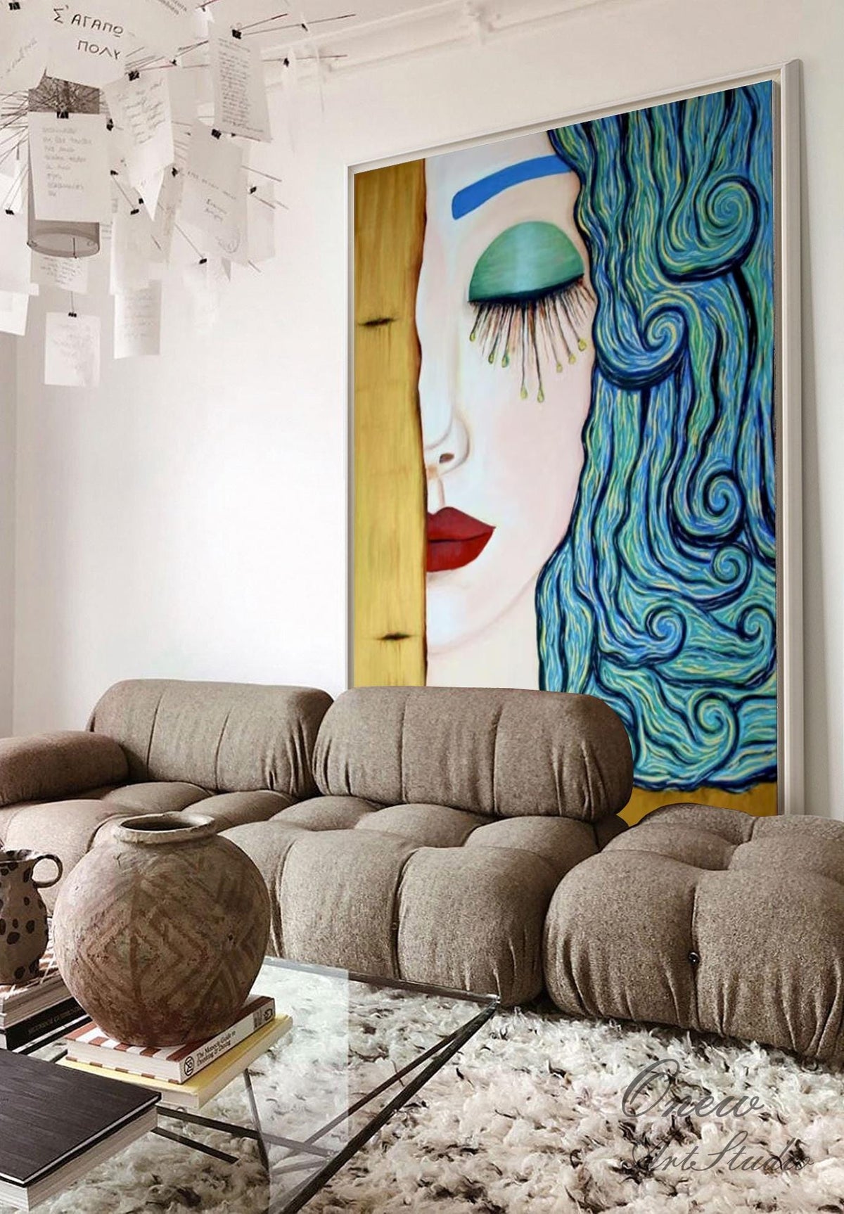 May include: A large framed painting of a woman's face with blue hair and a teal eye. The painting hangs above a modular brown sofa and a glass coffee table. A decorative vase sits on the table, and a white rug covers the floor.