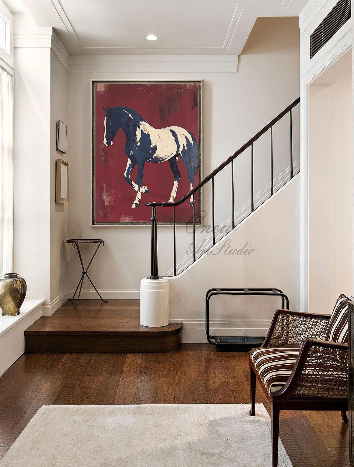 May include: Large framed painting of a horse in motion. The horse is depicted in white and navy blue against a deep red background. The artwork hangs on a white wall in a well-lit hallway with a staircase and a striped chair.
