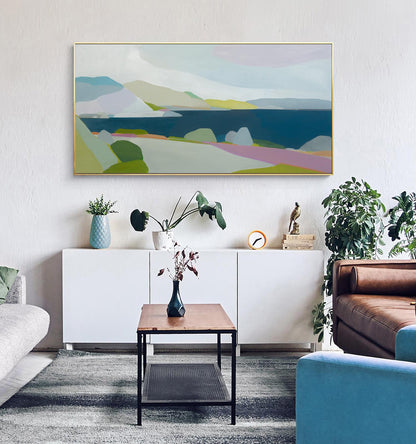 May include: An abstract landscape painting with a gold frame. The artwork features a coastal scene with blue water, green and pink land, and a light blue sky. The painting hangs above a white cabinet with decorative items.