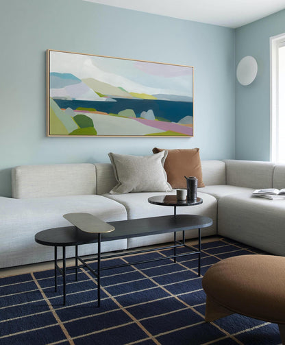 May include: A modern living room with a light blue wall and a large landscape painting. A white sectional sofa is adorned with throw pillows. A black coffee table with a gold accent sits on a navy blue grid patterned rug. A brown ottoman is in the foreground.