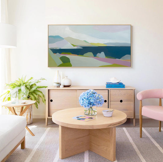 May include: A living room scene featuring a large landscape painting with blue, green, and purple hues. A wooden cabinet, round coffee table, and pink chair are in the room. A vase of blue hydrangeas sits on the table.
