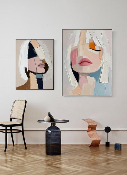 May include: Two abstract portrait paintings. The artwork features faces with bold brushstrokes and a muted color palette of pink, beige, and blue. The paintings are framed and displayed on a white wall.
