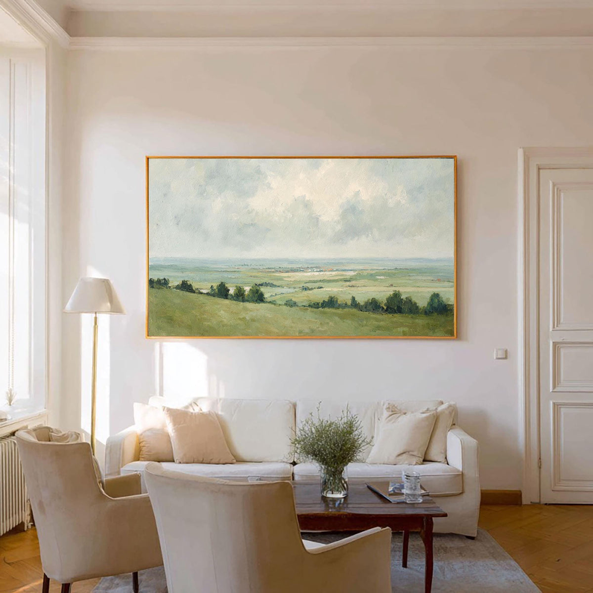 May include: A landscape painting with a gold frame hangs above a white sofa. The artwork depicts a green field under a cloudy sky. Two beige armchairs and a floor lamp are in front of a white wall.