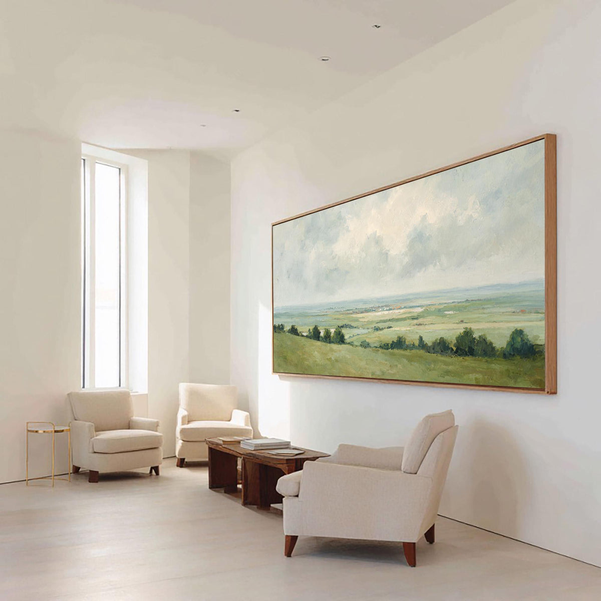 May include: A large landscape painting in a wooden frame hangs on a white wall. The painting depicts a green and yellow field under a cloudy sky. Three cream-colored armchairs and a wooden coffee table are in front of the painting.