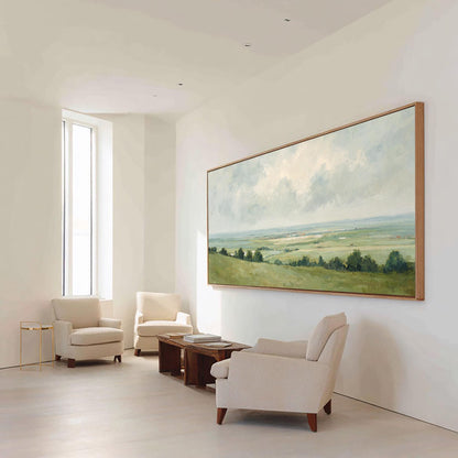 May include: A large landscape painting in a wooden frame hangs on a white wall. The painting depicts a green and yellow field under a cloudy sky. Three cream-colored armchairs and a wooden coffee table are in front of the painting.