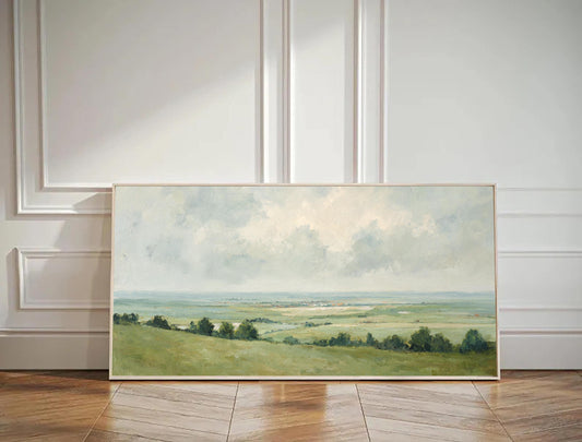May include: Landscape painting in a light wooden frame. The artwork depicts a serene vista of rolling green hills, a distant body of water, and a cloudy sky. The color palette is soft, with muted greens, blues, and creams.