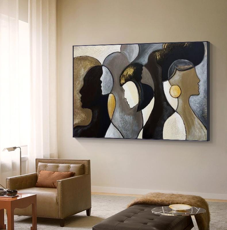 May include: Abstract art canvas featuring silhouettes of faces in shades of black, gold, and gray. The artwork is displayed in a living room setting, with a brown leather armchair and a small side table visible in the foreground.