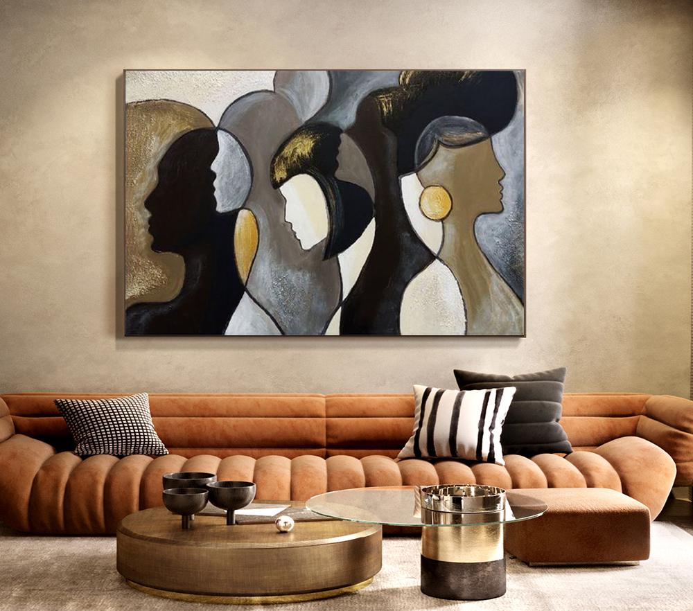 May include: Abstract wall art featuring three stylized female profiles in shades of black, gold, and gray. The artwork is displayed above a brown sofa with decorative pillows and a round coffee table.