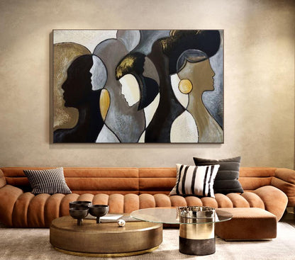 May include: Abstract wall art featuring three stylized female profiles in shades of black, gold, and gray. The artwork is displayed above a brown sofa with decorative pillows and a round coffee table.