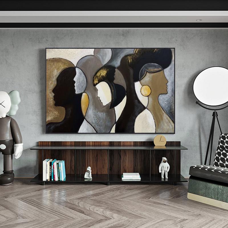 May include: Abstract painting featuring silhouettes of diverse faces in shades of black, gold, and beige. The artwork hangs above a dark wood and glass cabinet, with decorative items and books. The room has a modern aesthetic with a floor lamp and patterned chair.