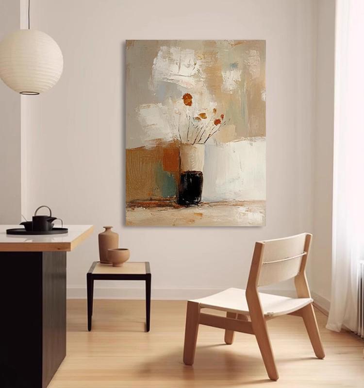 May include: Abstract art painting featuring a vase with dried flowers. The vase is black and cream, set against a backdrop of neutral tones including beige, white, and brown. The artwork is displayed in a well-lit room with a modern chair.