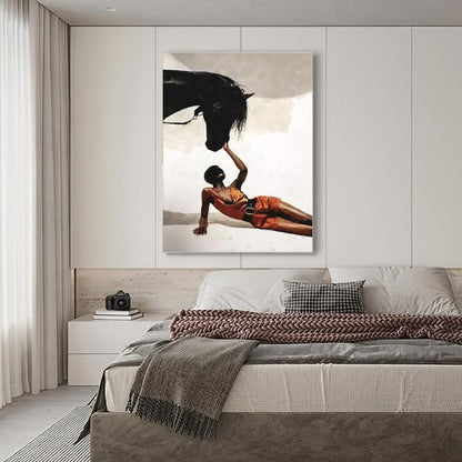 May include: A framed art print depicts a person in orange clothing interacting with a black horse. The person is lying down, reaching up to touch the horse's head. The background is a neutral color, and the artwork is displayed above a bed.