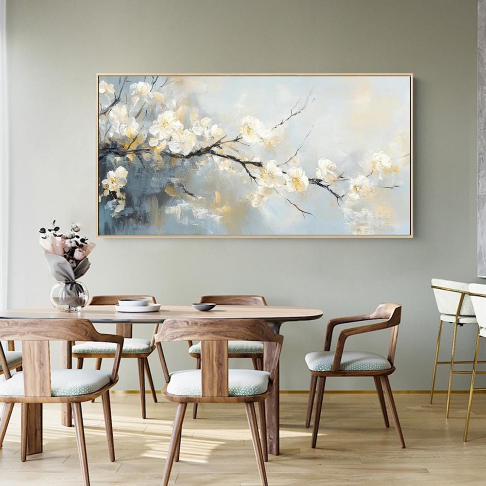 May include: A framed floral painting featuring white blossoms on a dark branch against a soft blue and gold background. The artwork hangs above a wooden dining table with matching chairs, creating a serene and elegant atmosphere.