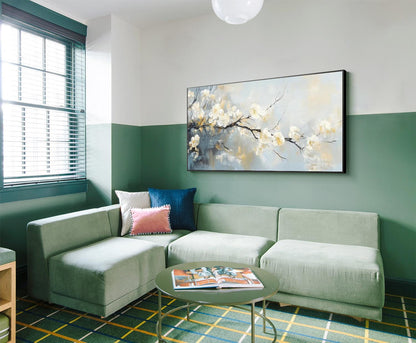 May include: A modern living room with a sage green sectional sofa, a round green coffee table, and a large framed painting of white flowers on a branch. The room has a green and white color scheme with a patterned rug.