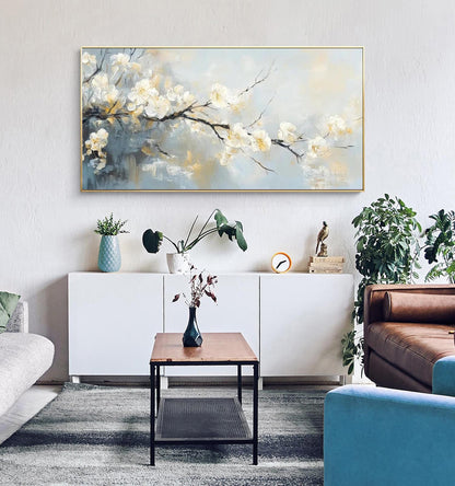 May include: An abstract painting of white blossoms on a branch against a soft blue and yellow background, framed in gold. The artwork hangs above a white cabinet, with a coffee table and sofa in the foreground. The room is decorated with plants and a rug.