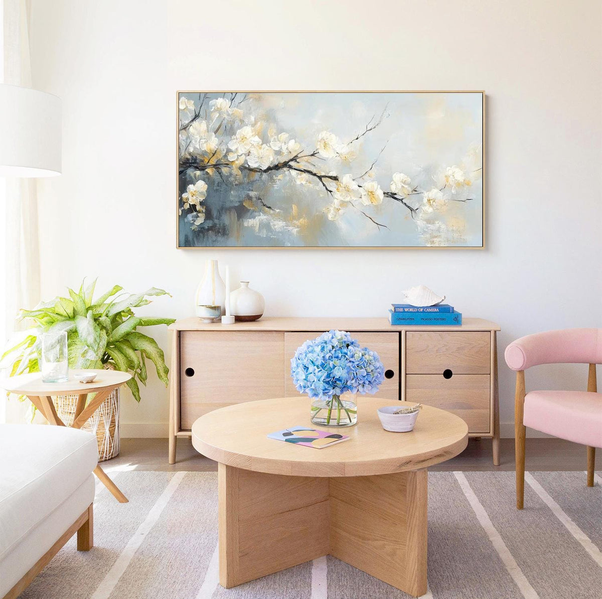 May include: A living room scene featuring a large framed painting of white blossoms on a branch against a blue and gray background. A wooden cabinet, round coffee table, and pink chair are also visible, creating a modern and inviting space.