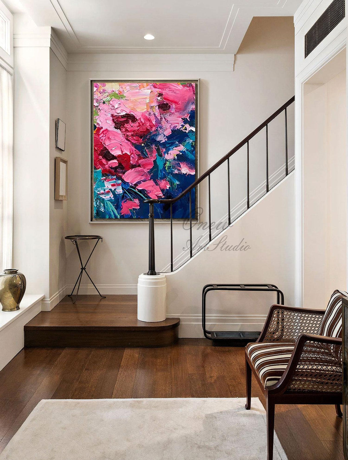 May include: Large abstract floral painting with vibrant pink, red, and blue brushstrokes. The artwork is framed and hangs on a white wall above a dark wooden staircase. The room features a chair and a decorative stand.