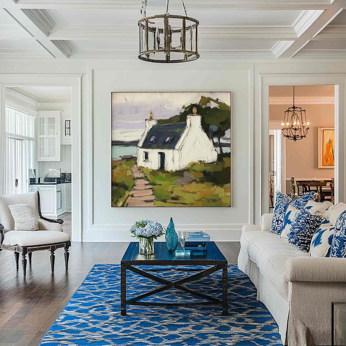 May include: A large square painting of a white cottage with a dark roof sits on a white wall above a dark blue wooden coffee table. The room features a blue and white patterned area rug, a white sofa with blue pillows, and a white armchair.