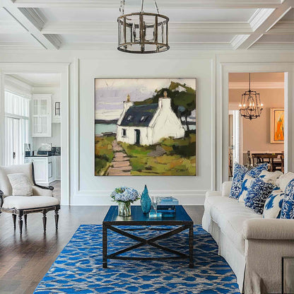 May include: A large square painting of a white cottage with a dark roof sits on a white wall above a dark blue wooden coffee table. The room features a blue and white patterned area rug, a white sofa with blue pillows, and a white armchair.