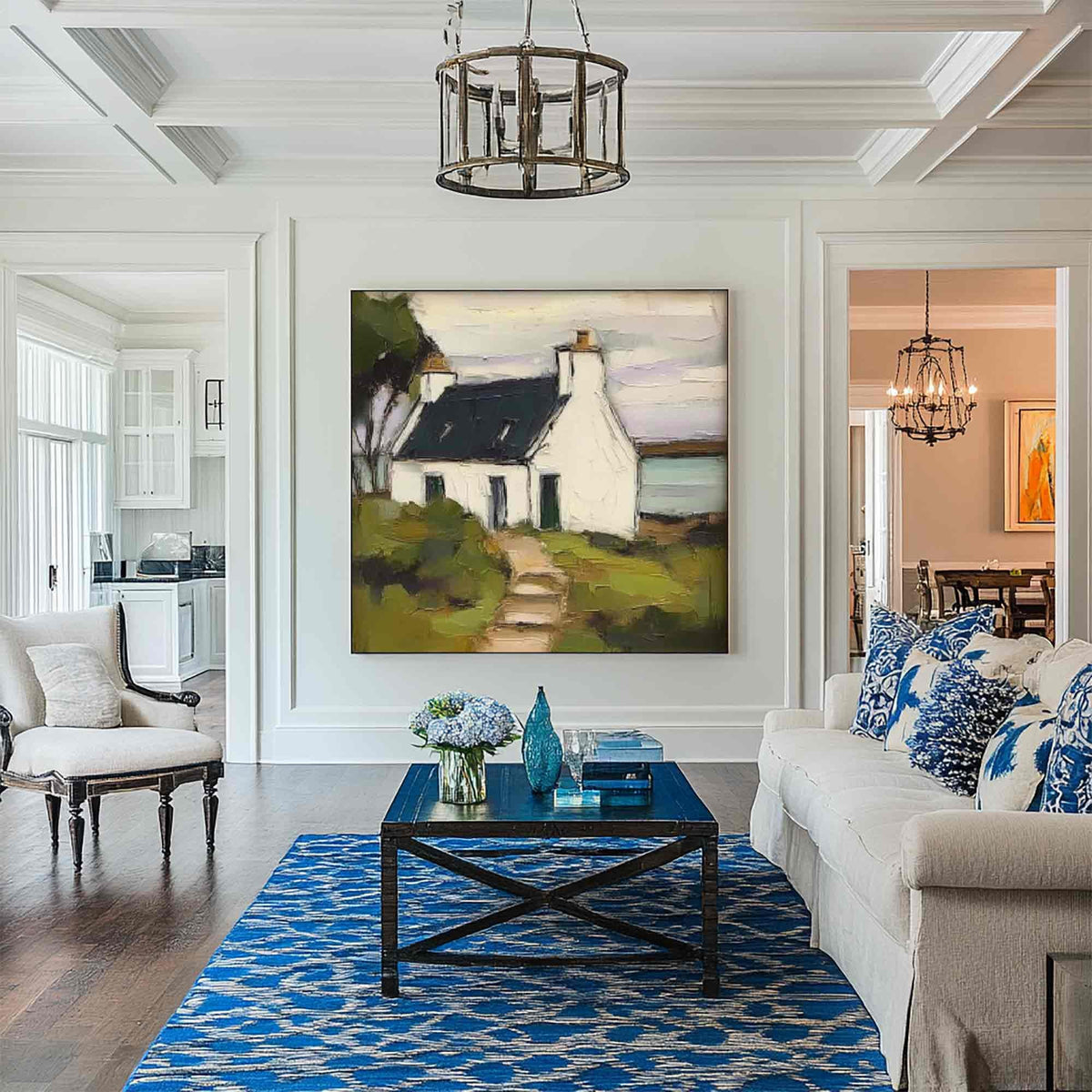May include: A large square oil painting depicting a white cottage with a dark roof and a path, displayed on a white wall above a dark blue wooden coffee table. The artwork features muted tones of green, brown, and white in a landscape setting.