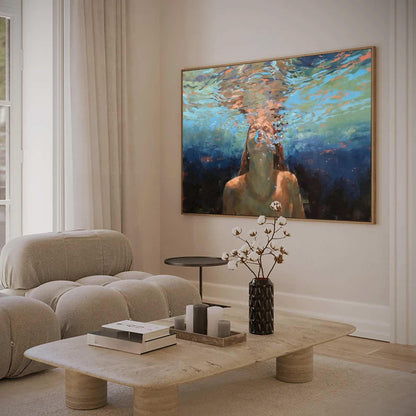 May include: A large framed painting depicts a person underwater, with bubbles rising towards the surface. The artwork is displayed above a stone coffee table with books, candles, and a black vase with cotton stems. A beige sofa and white curtains are also visible.