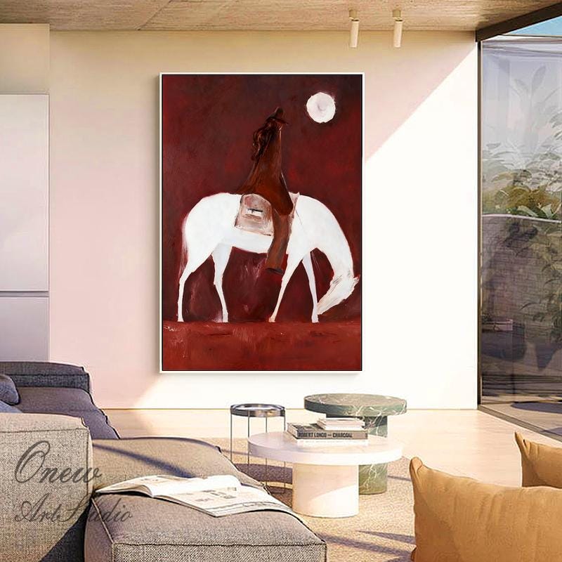 May include: An abstract painting depicts a rider on a white horse against a deep red background. The rider is clad in brown, and a white moon is in the upper right. The artwork is framed and displayed in a modern living space.