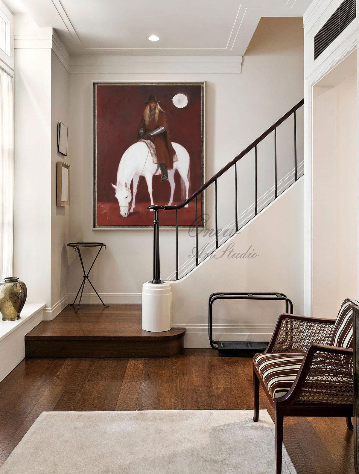 May include: An oil painting depicts a person riding a white horse under a full moon against a deep red background. The artwork hangs on a white wall above a dark wood staircase. A striped chair and a decorative table are in the foreground.
