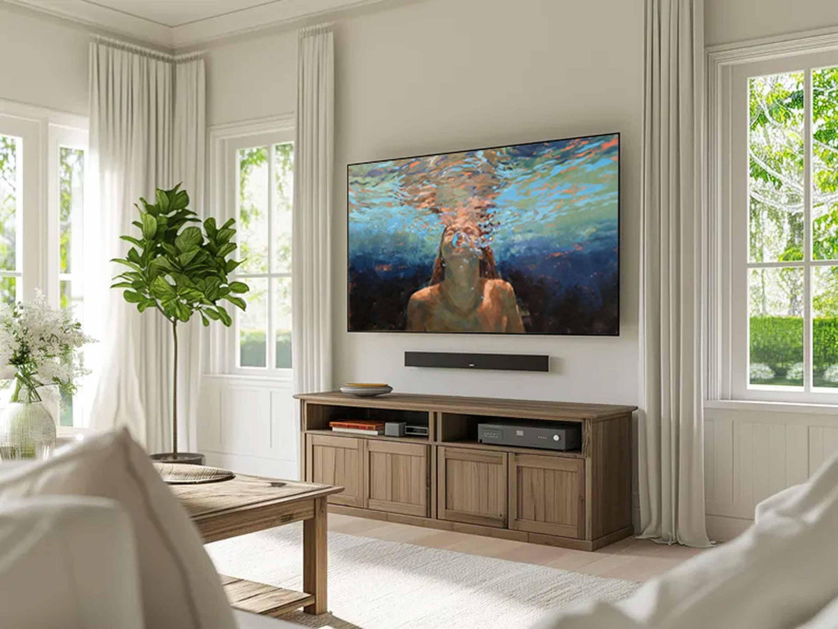 May include: A living room with a large flat-screen TV mounted on a white wall. Below the TV is a wooden cabinet with doors and open shelves. A soundbar is mounted below the TV. The room has large windows with white curtains and a green plant.