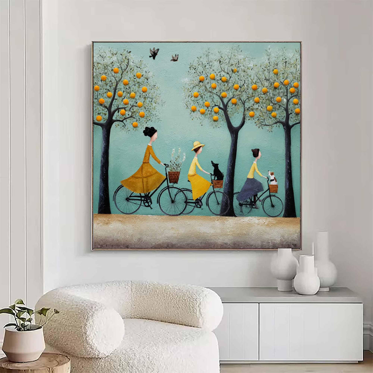 May include: A framed painting depicts three figures riding bicycles beneath orange trees. Each person wears a dress and has a basket on their bike, with a dog in one basket. The background is a light blue with birds flying above.