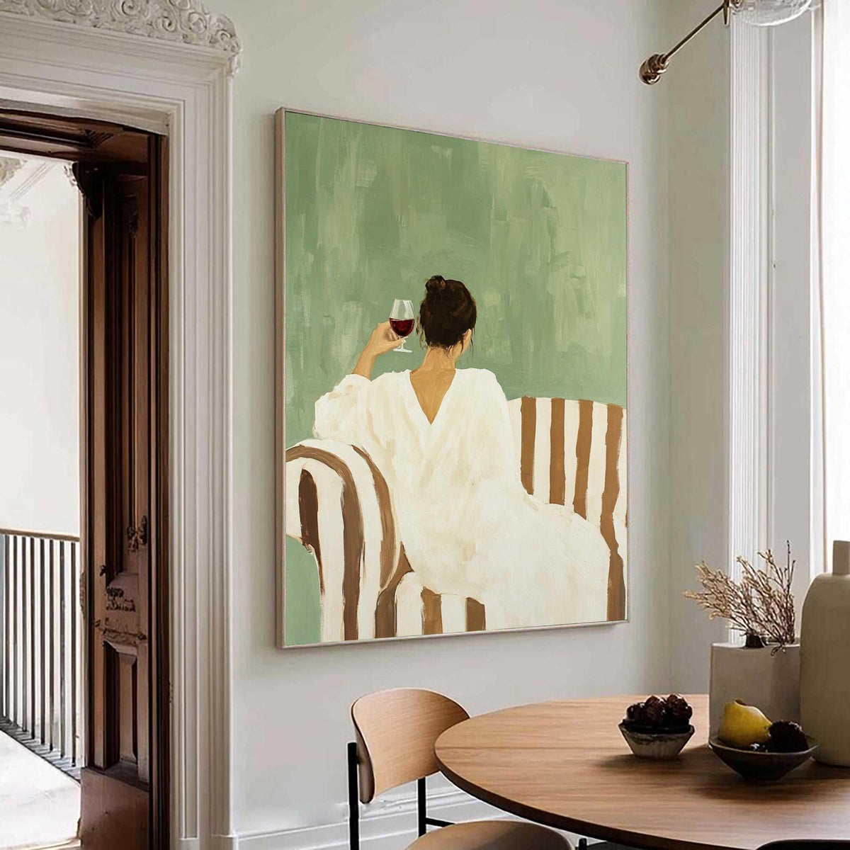 May include: An abstract painting depicts a person in a white dress seated on a striped sofa, holding a glass of red wine. The background is a muted green, and the artwork is framed in a light-colored wood.