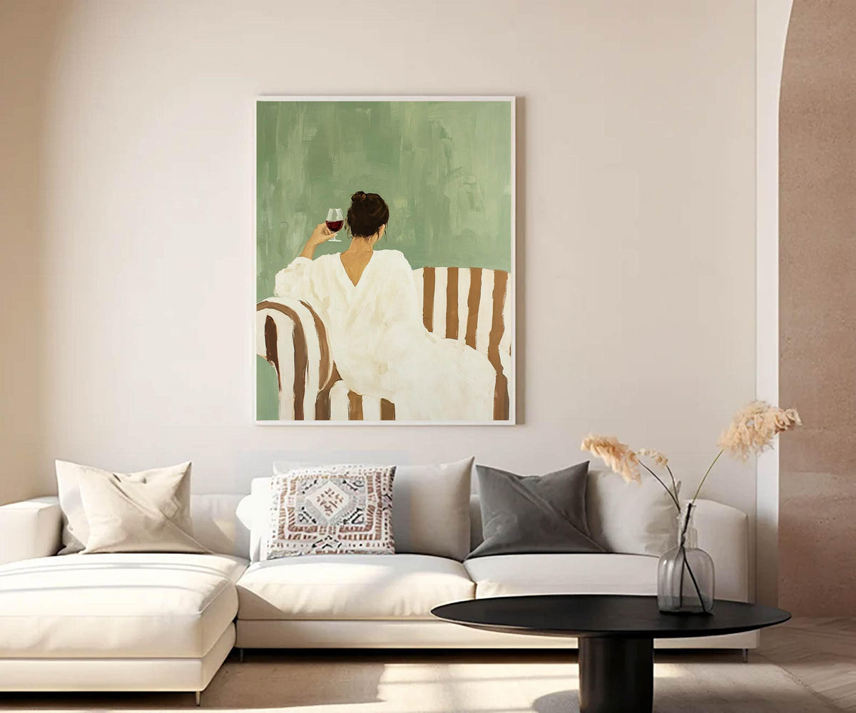 May include: An art print depicts a person in a white dress, seated on a striped sofa, holding a glass of red wine. The background is a muted green, and the artwork is framed in white. The scene evokes a sense of relaxation and leisure.