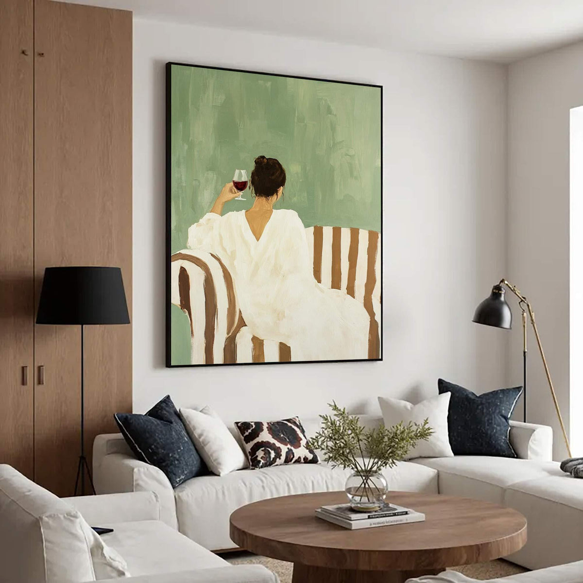 May include: An abstract painting depicts a person in a white dress seated on a striped sofa, holding a glass of red wine. The background is a muted green, and the artwork is framed in black. The painting hangs above a white sofa with decorative pillows.