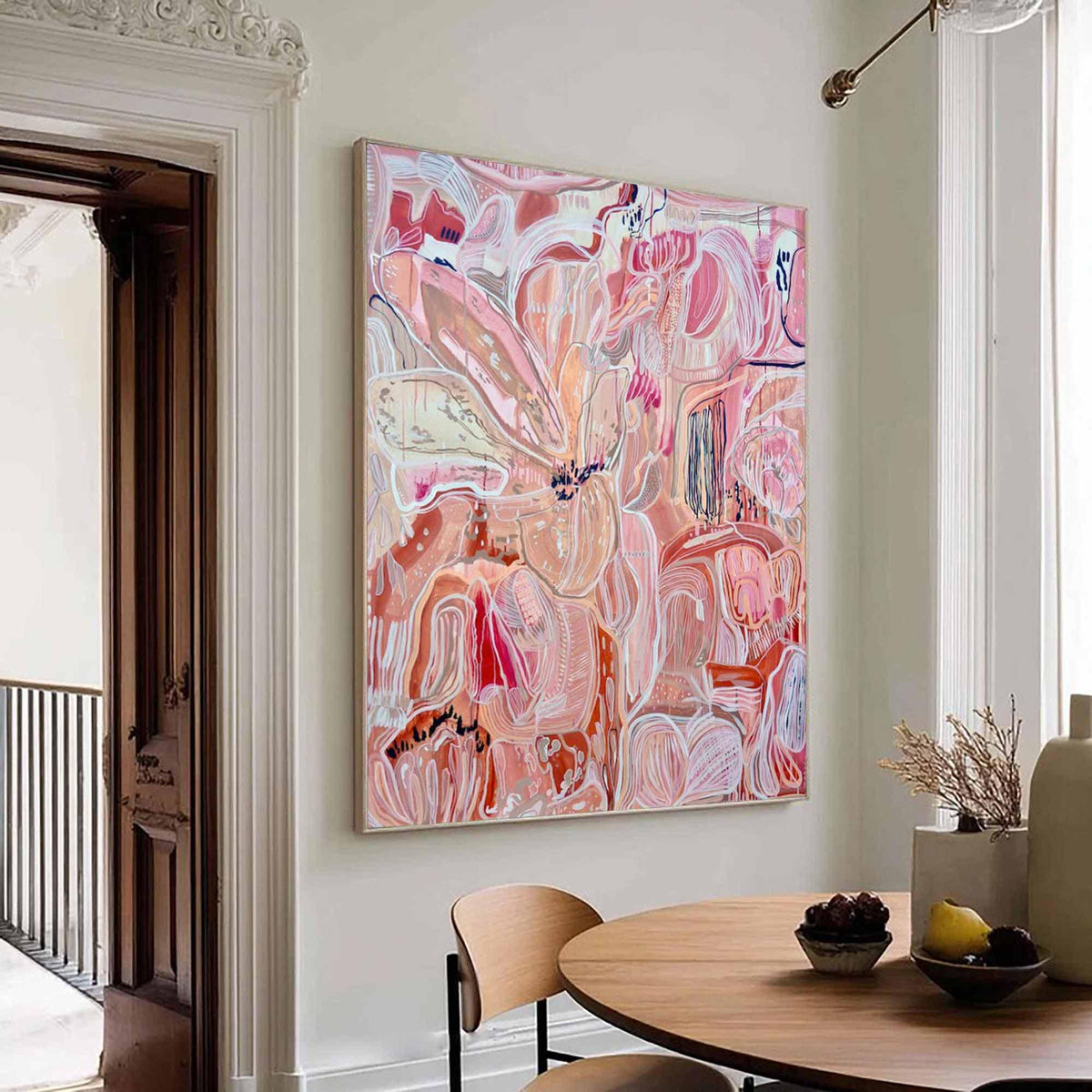 May include: Large abstract painting with a floral theme in shades of pink, red, and white. The artwork is displayed on a wall in a room with a wooden table and chairs. The painting has a light wooden frame.