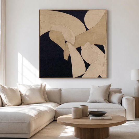 May include: Abstract art piece with a dark navy background and beige and off-white organic shapes. The artwork is framed in a light wood tone and hangs above a beige sectional sofa. The room is well-lit with natural light.