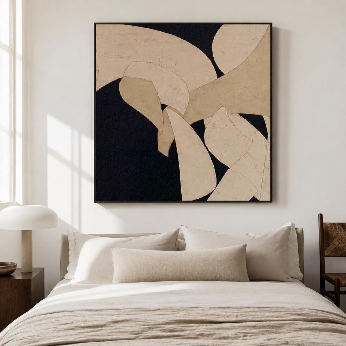 May include: Abstract art piece with a black background and beige, curved shapes. The artwork is framed in a black border and hangs above a bed with beige bedding and pillows. A wooden side table and lamp are visible.