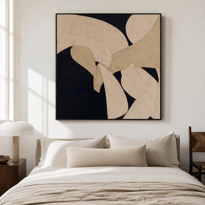 May include: Abstract art piece with a black background and beige, curved shapes. The artwork is framed in a black border and hangs above a bed with beige bedding and pillows. A wooden side table and lamp are visible.
